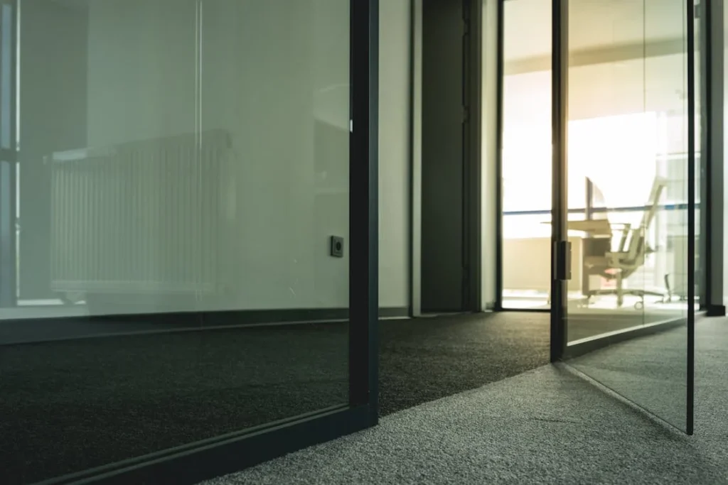 Modern office interior with floor-to-ceiling glass windows allowing natural light to illuminate workspace with office chairs and carpeted flooring. Clean windows and office productivity.