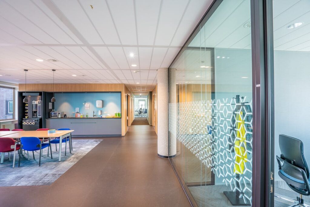 Modern office space with floor-to-ceiling glass walls, geometric patterned glass panels, open kitchen area, and contemporary furniture showcasing professional business environment with clean windows serviced by Brightness window cleaning